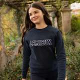LeMieux AW26 Young Rider Shay Varsity Crew Varient Navy. A young woman with long brown hair stands outdoors wearing a dark navy crewneck sweatshirt with 'LEMIEUX' printed across the chest in white outlined block letters, paired with matching dark pants.