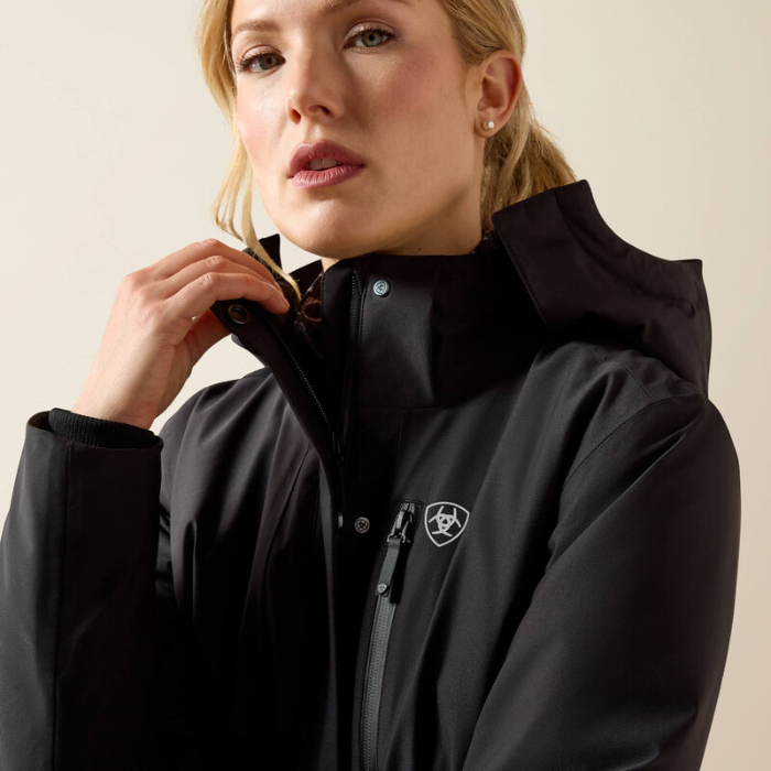 A close-up of a woman wearing a black hooded jacket with a high collar and a small shield logo on the chest, photographed against a plain light beige studio background.