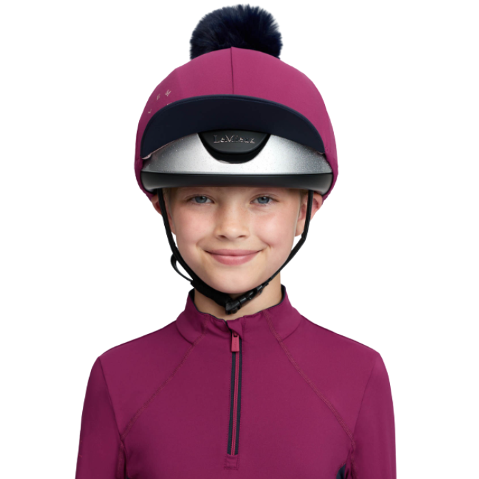 LeMieux AW26 Classique Pom Hat Silk Variant Colour Blackberry.
A child wearing a magenta riding helmet with a black pom-pom and visor, paired with a matching magenta quarter-zip top, against a plain white background.