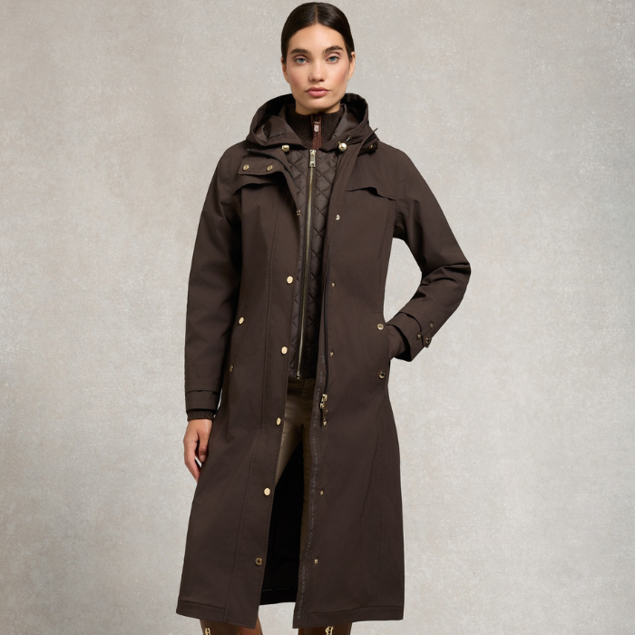 Holland Cooper Folgate Rain Coat Variant Chocolate. A woman modeling a long dark brown hooded parka coat with gold-tone snap buttons, shown against a textured light gray studio background.