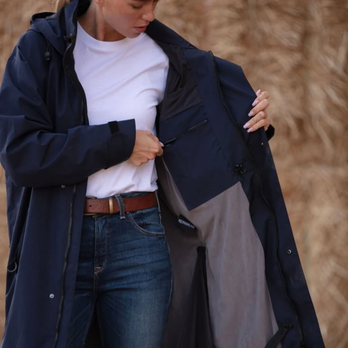 A woman wearing a navy blue hooded jacket over a white t-shirt and blue jeans, opening the jacket to show the gray interior lining, set against a background of dry grass.