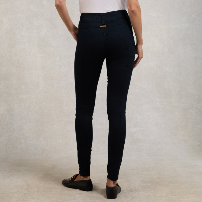 Holland Cooper Jodhpur Jean variant Ink Navy. Rear view of a model wearing dark navy blue skinny jeans with a small gold brand tab on the back waistband, paired with a white top and black loafers, against a textured beige studio backdrop.