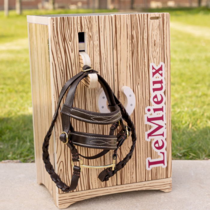 LeMieux AW26 Toy Pony Tack Locker Wooden Varienf Brown. Small wooden tack box with toy pony bridle hung outisde with LeMieux printed on the door on a concrete floor with grass garden behind. 