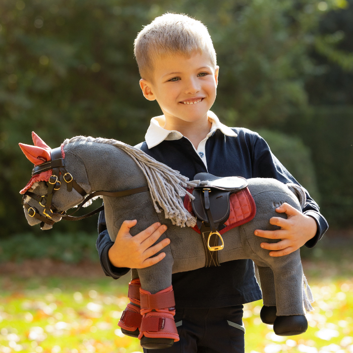 LeMieux AW26 Toy Pony Variant Colour Smokey. Young person wearing navy top holding a pplush toy grey pony on faded garden background.