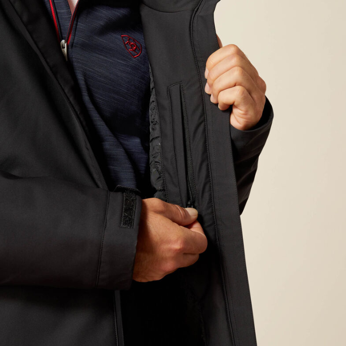 Close-up of a person wearing a black zip-front jacket and a navy knit top, opening the jacket to show the inner pocket and interior lining. A small red embroidered logo is visible on the chest.