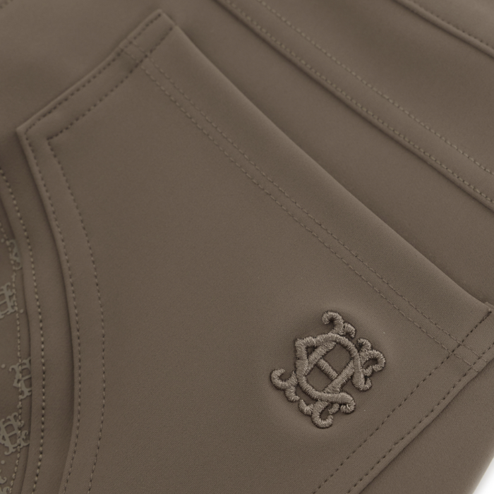 Holland Cooper Chiltern Contour Breeches Variant Colour Mocha.
Close-up of a taupe-brown leather or leather-look garment, showing a stitched pocket with an embroidered monogram logo.