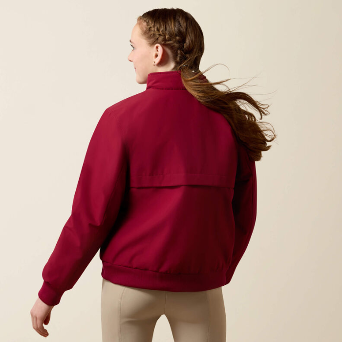 A woman shown from the back wearing a dark red or burgundy zip-front jacket with a high collar and beige pants, photographed against a plain light beige studio background.
