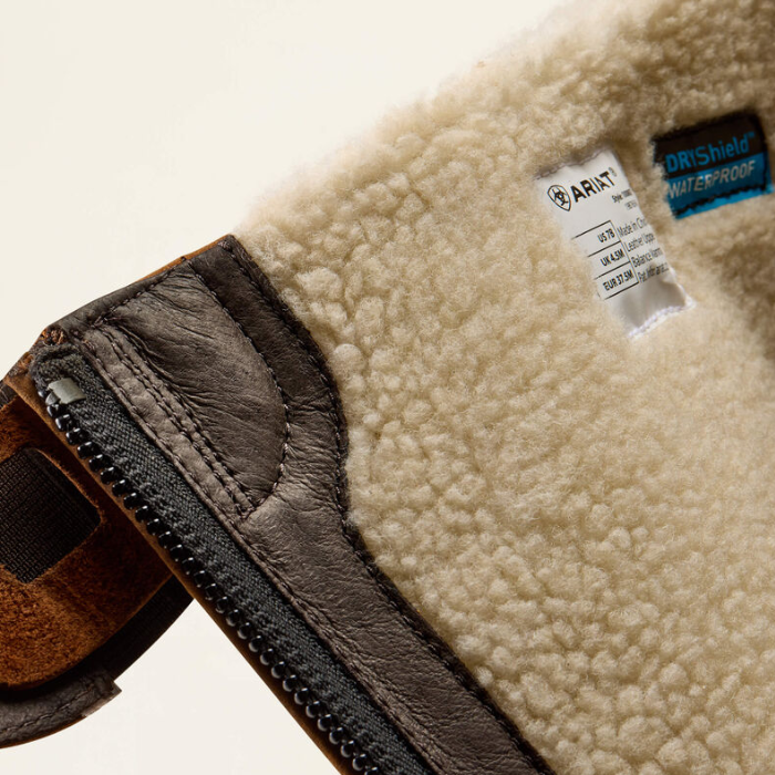 Close-up detail of a boot showing a cream sherpa fleece lining, a dark brown leather zipper flap, and a white ARIAT brand tag alongside a blue DRiShield waters-proof label.