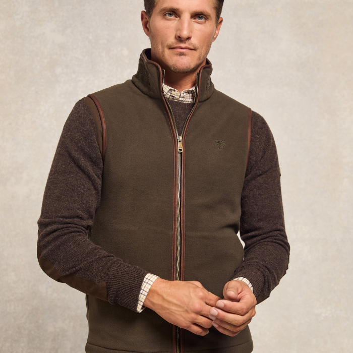 Holland Cooper Country Fleece Gilet (Men's) Variant Khaki. A man wearing a khaki zip-up fleece vest over a dark brown knit sweater and a patterned collared shirt, standing against a textured light gray background.