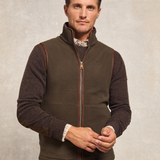 Holland Cooper Country Fleece Gilet (Men's) Variant Khaki. A man wearing a khaki zip-up fleece vest over a dark brown knit sweater and a patterned collared shirt, standing against a textured light gray background.