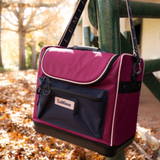 LeMieux AW26 Elite Pro Grooming Bag Varient Blackberry. A LeMieux shoulder bag in magenta and navy blue, hanging from a green fence post outdoors on a path covered with fallen autumn leaves.