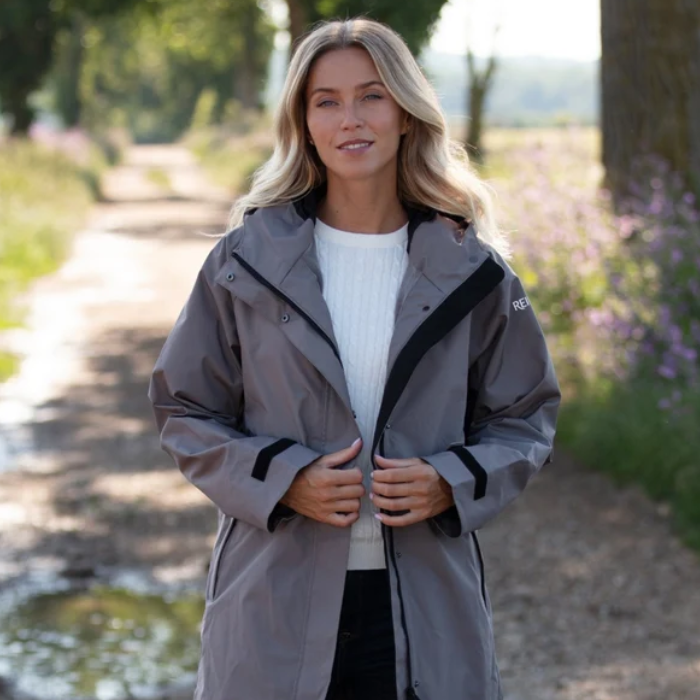 Reincoat Parka Lite Varient Dune. A woman wearing a gray waterproof jacket with black trim stands on a country path outdoors. The jacket has a high collar, front zipper, and black sleeve cuffs.