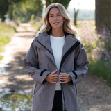 Reincoat Parka Lite Varient Dune. A woman wearing a gray waterproof jacket with black trim stands on a country path outdoors. The jacket has a high collar, front zipper, and black sleeve cuffs.
