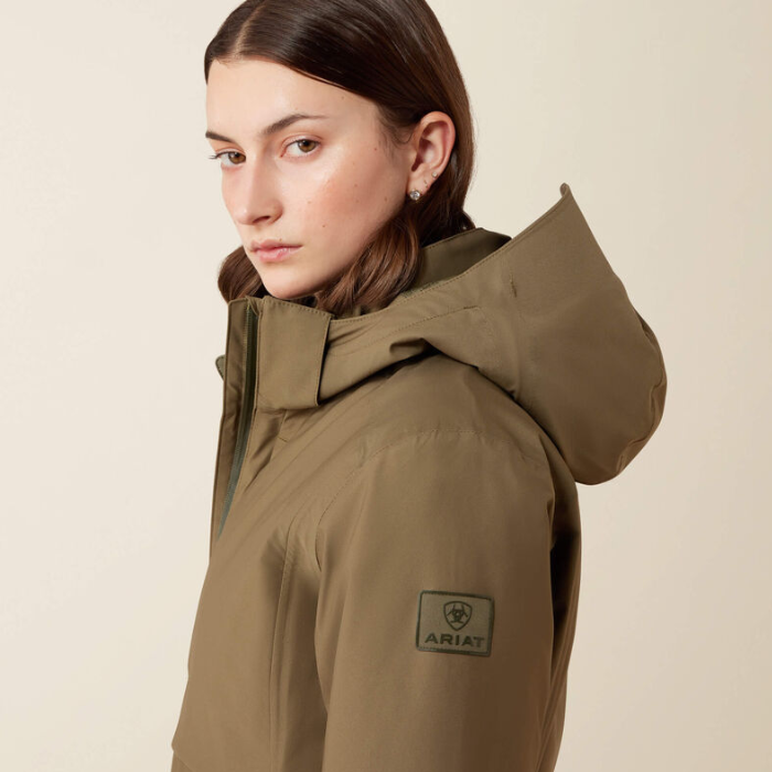 A woman wearing an olive green hooded jacket with a visible ARIAT logo patch on the sleeve, photographed against a plain light beige studio background.