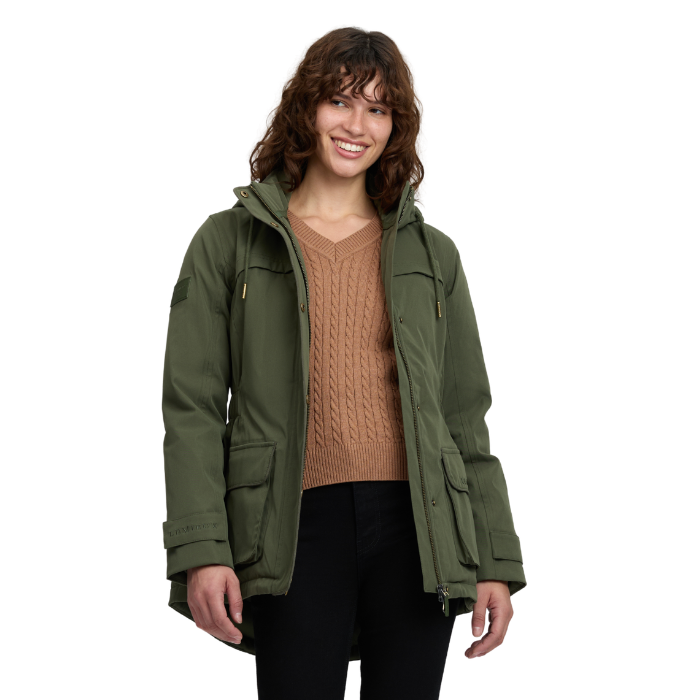 LeMieux AW26 Heritage Beatrice Waterproof Coat Variant Olive. A woman wearing an olive green hooded utility jacket over a tan cable-knit sweater and black pants, photographed against a plain white studio background.
