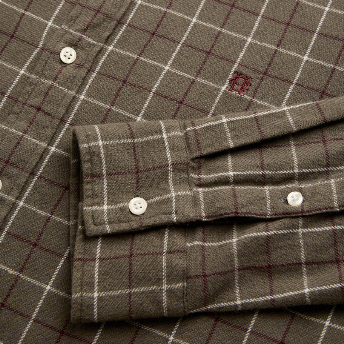 Holland Cooper Men's Classic Fit Brushed Cotton Shirt Variant Ruby Khaki Check. Close-up of a plaid button-up shirt fabric showing the cuff, white buttons, and a small embroidered logo on an olive green base with white and burgundy lines.