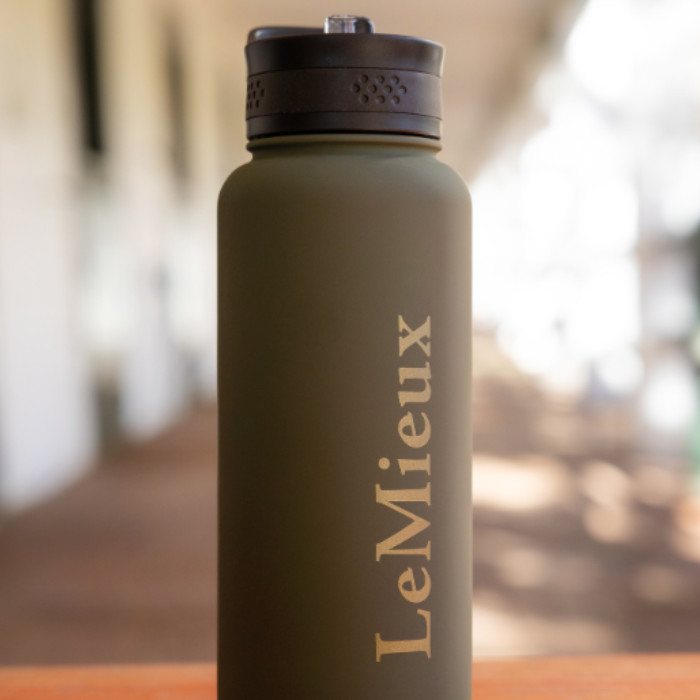 LeMieux AW26 Drinks Bottle Varient Willow Green. An olive green insulated water bottle with a black lid and the brand name 'LeMieux' printed vertically in gold, set against a blurred outdoor background.
