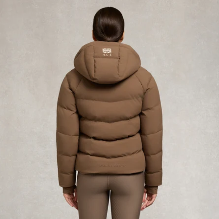 Holland Cooper Chiltern Yard Jacket Variant Chestnut. A woman viewed from the back wearing a brown hooded puffer jacket and matching brown leggings against a textured light gray studio background.
