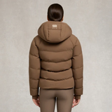 Holland Cooper Chiltern Yard Jacket Variant Chestnut. A woman viewed from the back wearing a brown hooded puffer jacket and matching brown leggings against a textured light gray studio background.