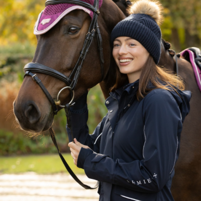 LeMieux AW26 Stormwear Performance Riding Coat Varient Navy. A woman smiling next to a brown horse outdoors, wearing a navy knit beanie with a beige pom-pom and a navy equestrian jacket with white 'LeMieux' text on the sleeve. The horse has a purple and pink blindfold.