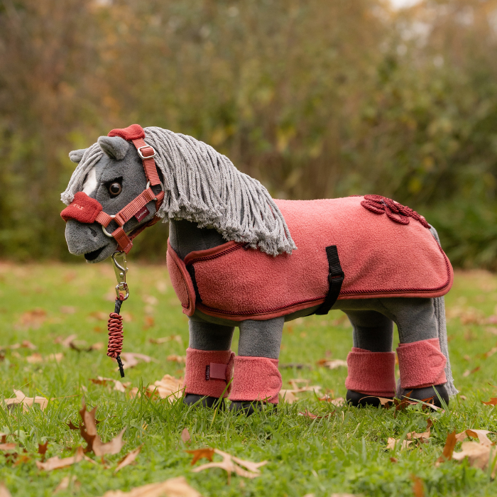 LeMieux AW26 Toy Pony Bandages Variant Colour Spiced Earth. A plush toy pony wearing an equestrian costume including tack, pad and bandages on faded garden 