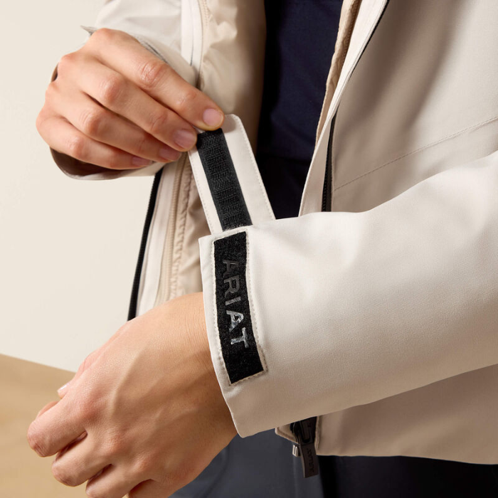 Close-up of a person wearing a light beige jacket and pulling the zipper tab, which features a black strap with the brand name 'ARIAT' embroidered vertically in grey.