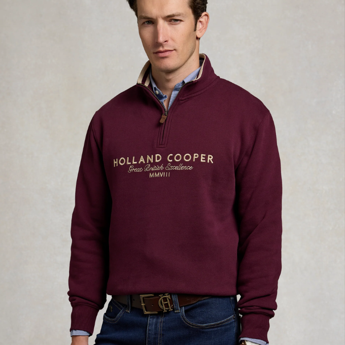 Holland Cooper Heritage Quarter Zip Sweat (Men's) Variant Mulberry. Man wearing a Mulberry 1/4 zip sweatshirt with Holland Cooper branding across the chest in white agaisnt a grey background. 