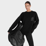 Woman holding a black leather saddle with a plain background wearing a black jumper
