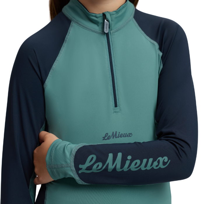 LeMieux AW26 Mini Base Layer Varient Navy/Aloe. A close-up of a person wearing a long-sleeve quarter-zip athletic top in teal and navy blue, with the LeMieux logo printed on the chest and sleeve.