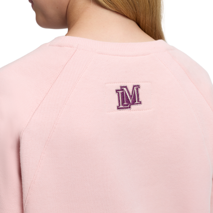 LeMieux AW26 Young Rider Shay Varsity Crew Varient Petal. Close-up back view of a person wearing a light pink crewneck sweatshirt with a purple embroidered 'LM' logo on the upper back.