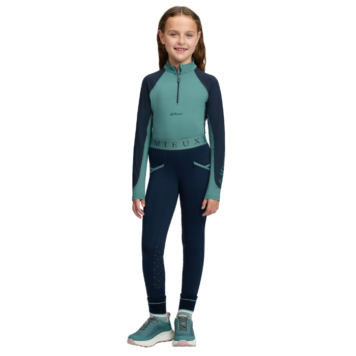LeMieux AW26 Mini Base Layer Varient Navy/Aloe. A young girl stands against a white background wearing a two-piece, featuring a teal and navy long-sleeve quarter-zip top and matching navy leggings, paired with teal and white sneakers.