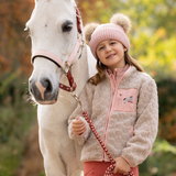 LeMieux AW26 Mini Thora Heart Fleece Varient Stone. A young girl stands next to a white horse outdoors, wearing a pink knit pom-pom beanie, a textured light beige fleece jacket with pink trim and a horse patch on the pocket, and pink pants. She is holding a pink and red braided lead rope.