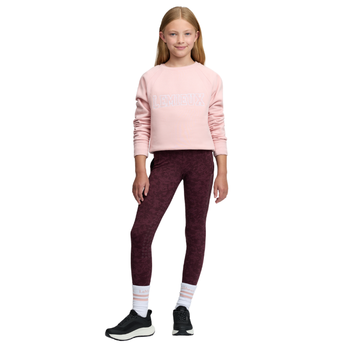 LeMieux AW26 Young Rider Shay Varsity Crew Varient Petal. A young girl modeling a light pink long-sleeve sweatshirt with 'LEMIEUX' printed in white across the chest, paired with maroon heathered leggings, white socks, and black sneakers, set against a plain white background.
