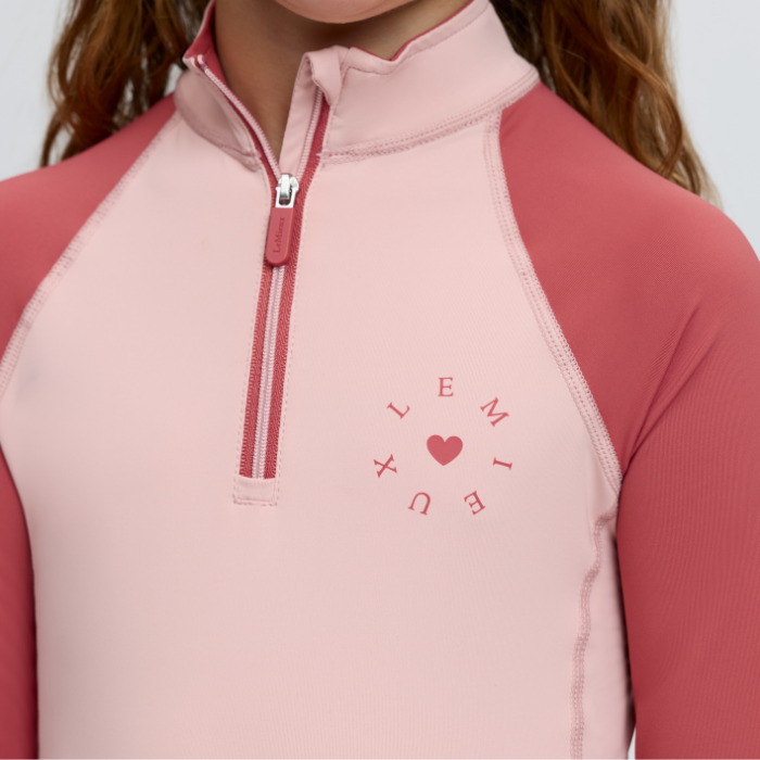 LeMieux AW26 Mini Base Layer Varient Petal. A close-up of a child wearing a light pink quarter-zip long-sleeve top with darker pink sleeves and a matching pink heart logo on the chest.