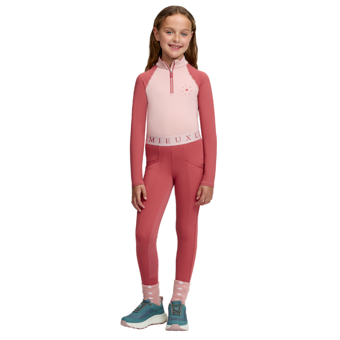 LeMieux AW26 Mini Base Layer Varient Petal. A young girl modeling a two-piece athletic set consisting of a long-sleeve quarter-zip top and matching leggings. The top is light pink on the front and darker pink on the sleeves and collar, with 'LeMieux' printed on the waistband.