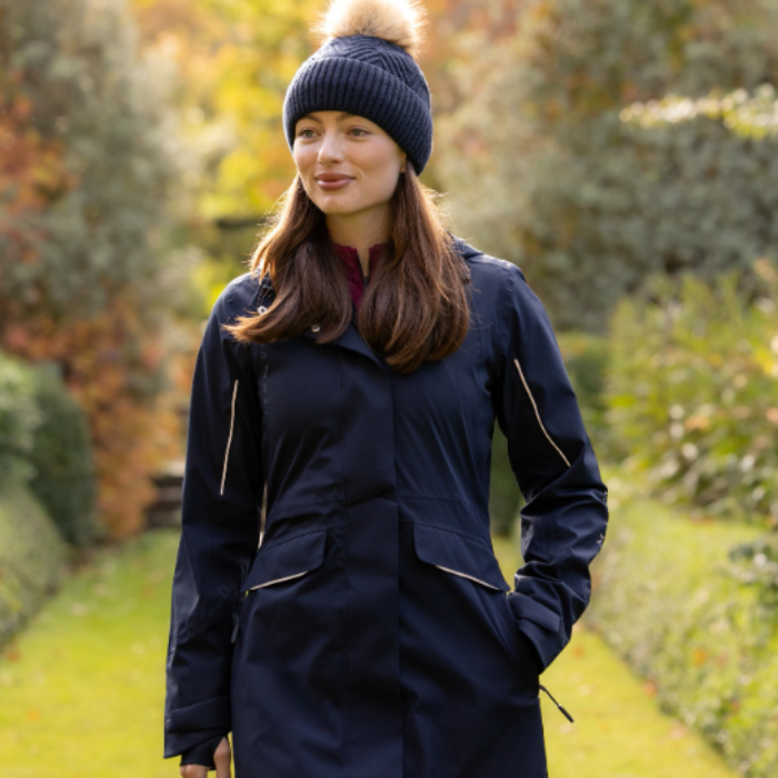 LeMieux AW26 Stormwear Performance Riding Coat Varient Navy. A woman wearing a navy blue waterproof coat with gold zipper accents and a matching navy blue knit beanie with a beige pom-pom, standing in a garden path.