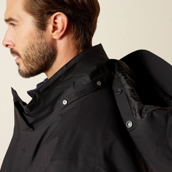 Close-up side profile of a man wearing a black waterproof jacket with a high collar and silver snap buttons, against a plain beige background.