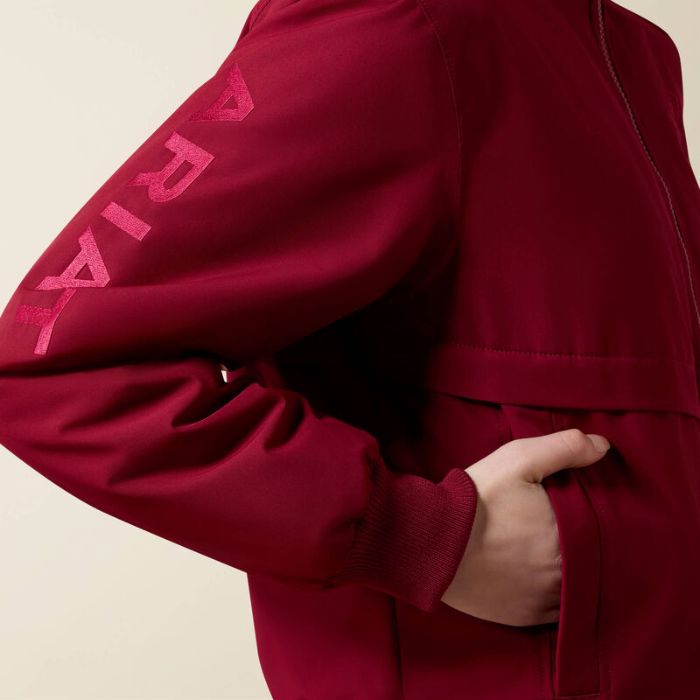 A close-up of a person wearing a deep red jacket, showing the sleeve with a pink 'ARIAT' logo and a hand tucked into a side pocket, against a light beige background.