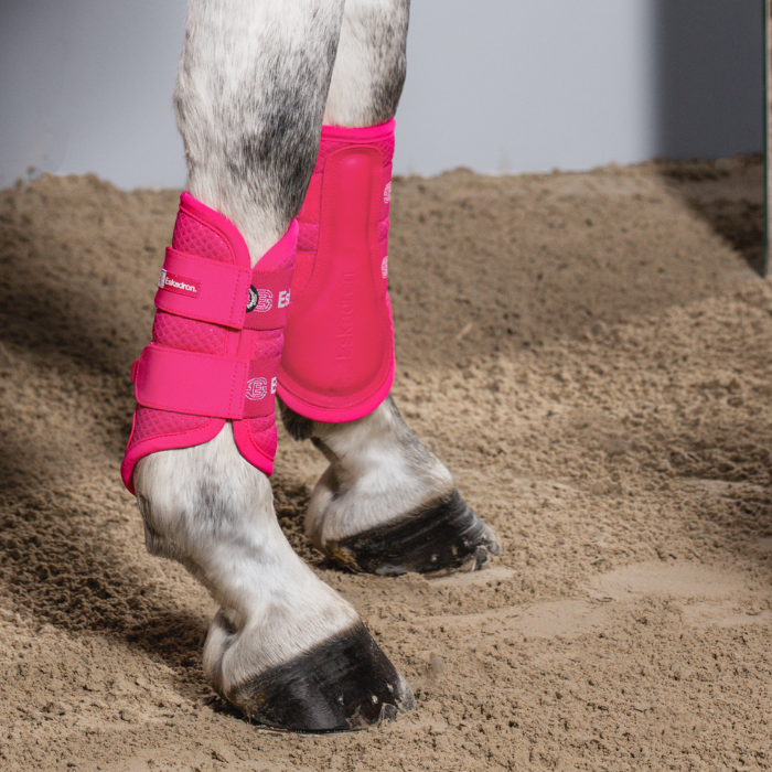 A pair of bright pink horse boots shown on a grey horses legs in a stable.