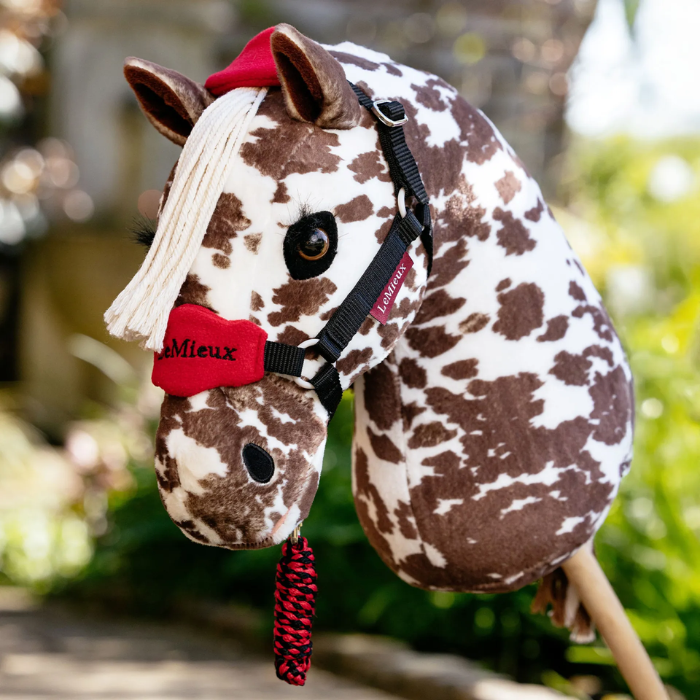 LeMieux AW26 Hobby Horse Vogue Headcollar & Leadrope Variant Colour Chilli. 
A plush toy horse head mounted on a stick, with a brown and white spotty body, white faux-fur mane, and chilli colour halter, photographed outdoors against a blurred garden background.