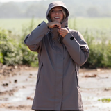 Reincoat Parka Lite Varient Dune. A woman wearing a hooded rain jacket stands outdoors in a muddy field, smiling and adjusting the hood. The jacket is a muted taupe-gray color.