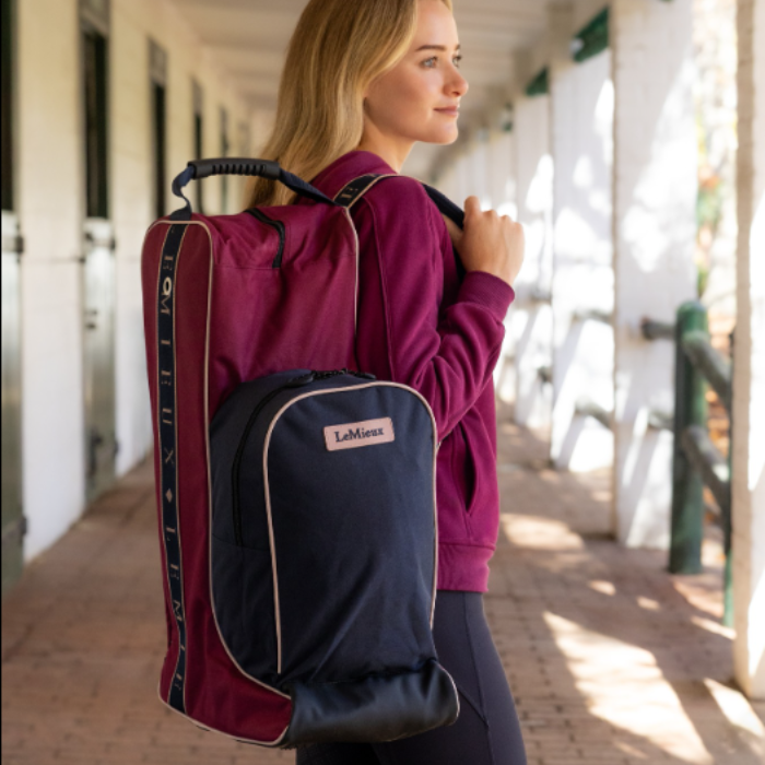 LeMieux AW26 Boot & Hat Bag Varient Blackberry. A woman in a maroon sweatshirt carries a large LaMieux hat and boot bag on her shoulder in an outdoor corridor with horse stables. 