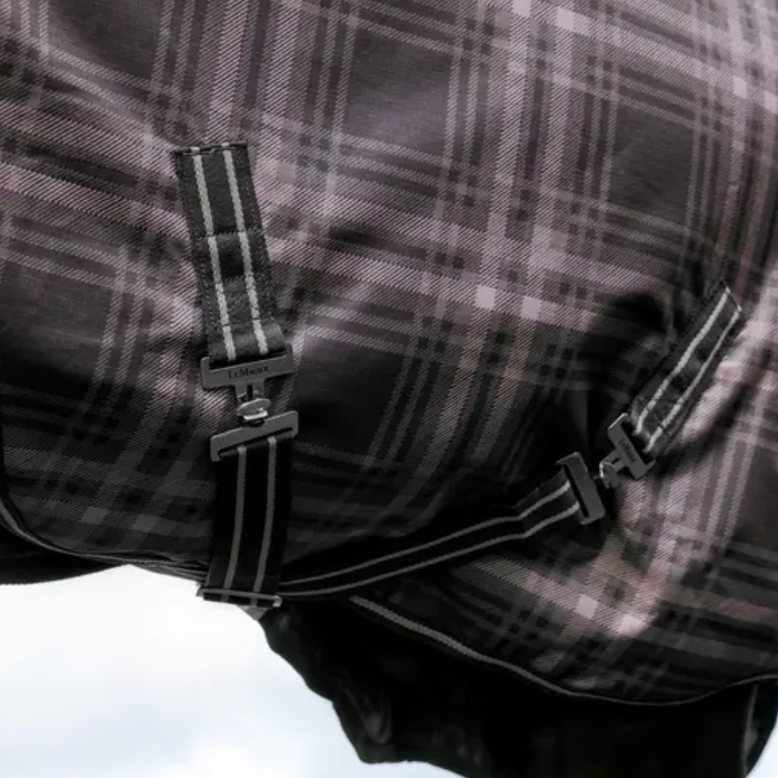 Close-up of a horse blanket with a black and gray plaid pattern, featuring adjustable straps with metal buckles and a visible LeMieux brand logo.