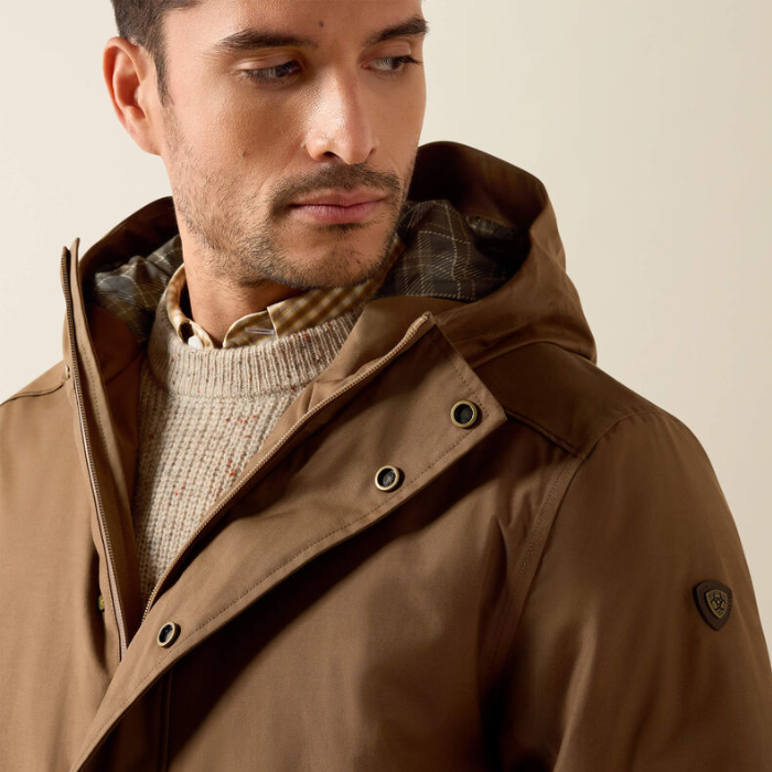 A close-up of a man wearing a brown hooded jacket over a beige knit sweater, photographed against a plain light beige background.