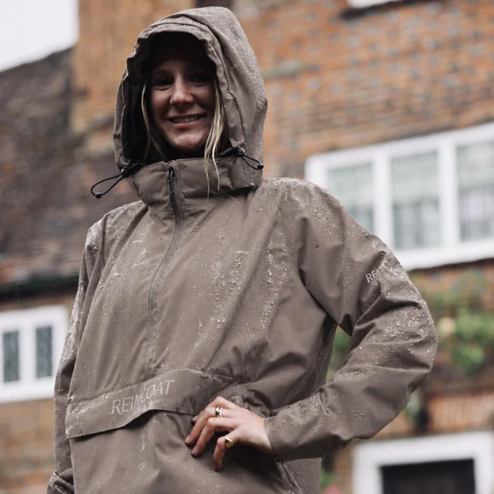Reincoat Smock Lite Varient Dune. Woman wearing a Dune coloured jacket with a kangaroo style pocket with "REINCOAT" branding in the rain. 