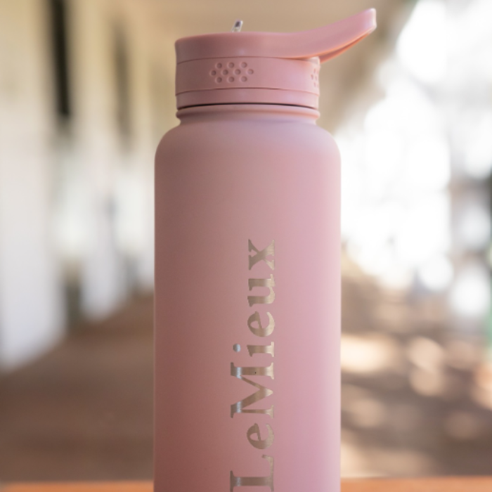 LeMieux AW26 Drinks Bottle Varient Wild Rose. A matte pink insulated water bottle with a flip-top lid, featuring the brand name 'LeMieux' printed vertically in a metallic gold tone, set against a blurred outdoor background.