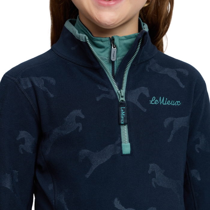 LeMieux AW26 Mini Maggie Horse Fleece Varient Navy. Close-up of a person wearing a navy quarter-zip fleece with a subtle horse pattern, teal inner collar, and teal zipper pull. The brand name 'LeMieux' is embroidered on the chest.