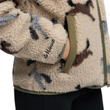 LeMieux AW26 Mini Theo Pony Fleece Varient Willow Green. Close-up of a person wearing a cream sherpa fleece jacket with a brown and gray animal print pattern, olive green trim, and a zippered pocket. The brand name 'LeMieux' is embroidered on the sleeve.