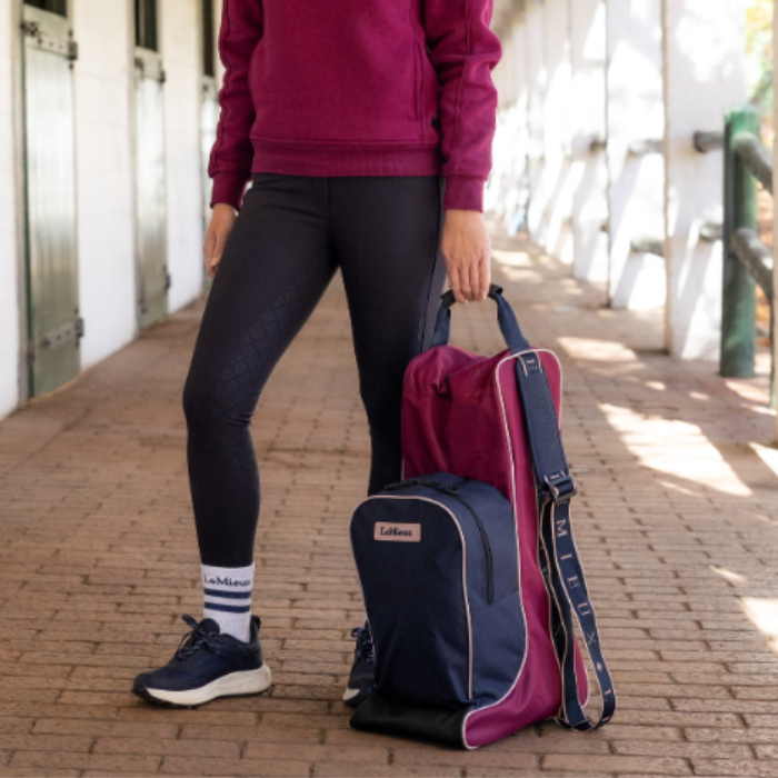 LeMieux AW26 Boot & Hat Bag Varient Blackberry. A person standing outdoors beside a brick walkway holding a LeMieux bag. The bag is a Blackberry and navy blue with pink piping and a white brand label.