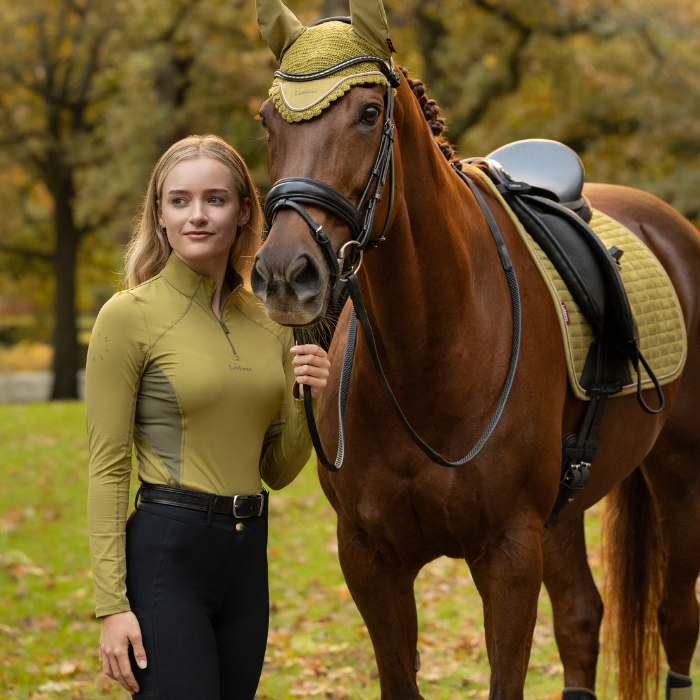LeMieux AW26 Classique Base Layer Variant Colour Gooseberry.
A woman in equestrian apparel stands beside a brown horse outdoors. She wears a fitted gooseberry green long-sleeve riding shirt and black riding pants, while the horse is outfitted with a matching olive-green and black bridle and saddle pad.